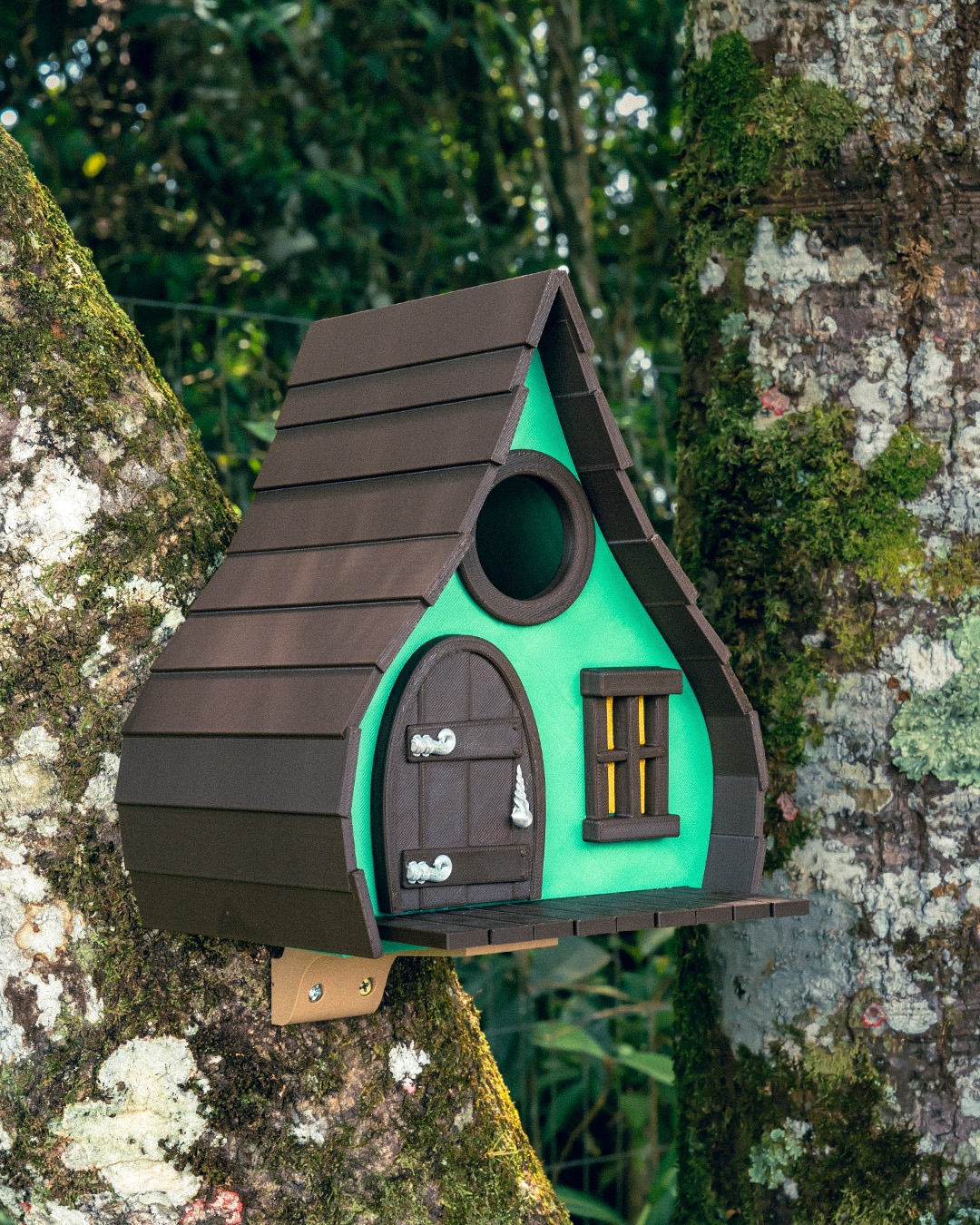 Colorful birdhouse designed like a small cottage, featuring a green and brown color scheme, set against a tree trunk in a lush forest environment. Ideal for attracting birds to gardens and outdoor spaces.