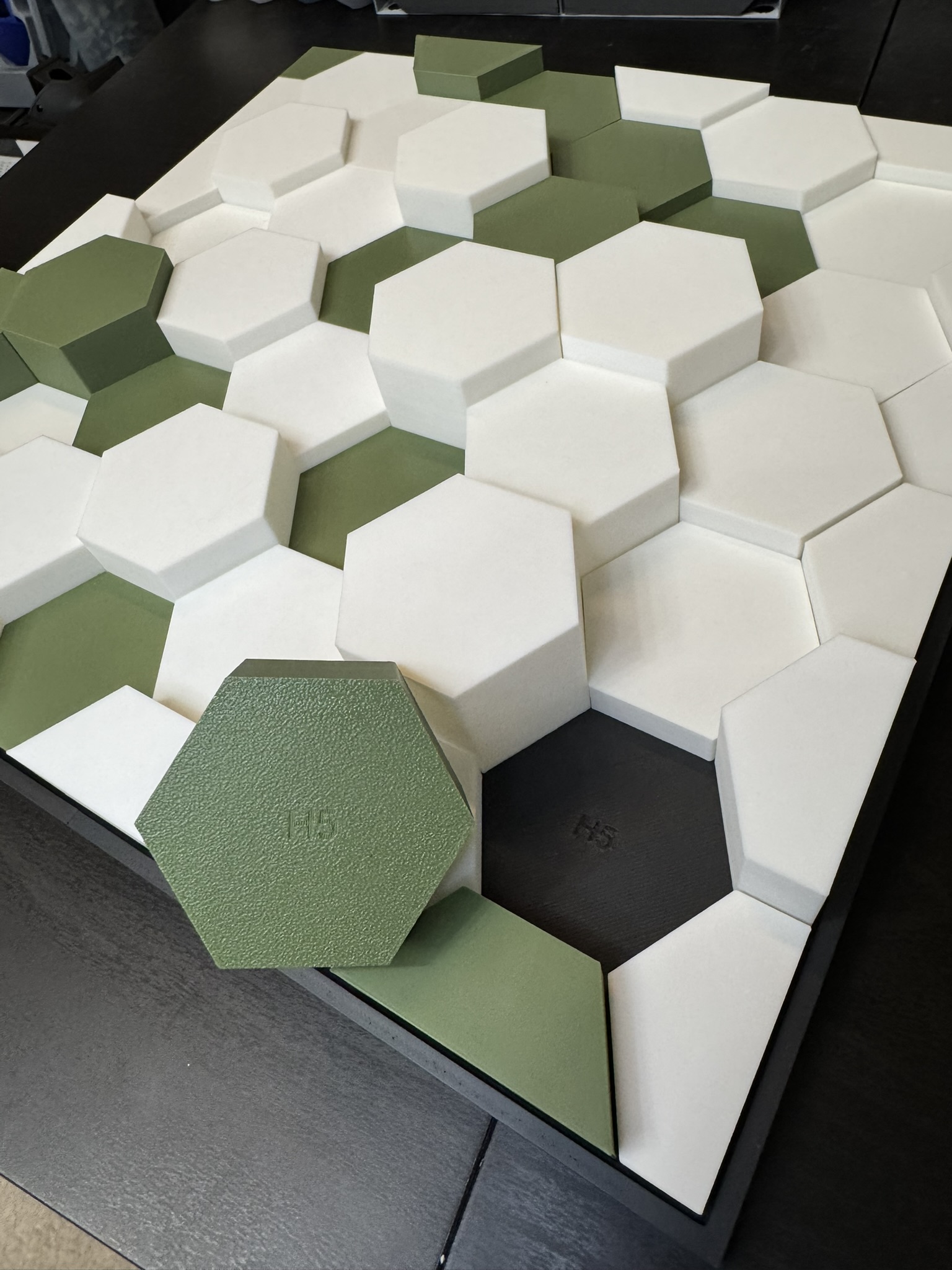 IMG_8494 Colorful hexagonal tiles in shades of green and white arranged on a black surface, showcasing a modern and geometric design. The image highlights a partially completed pattern with one green hexagon prominently placed.