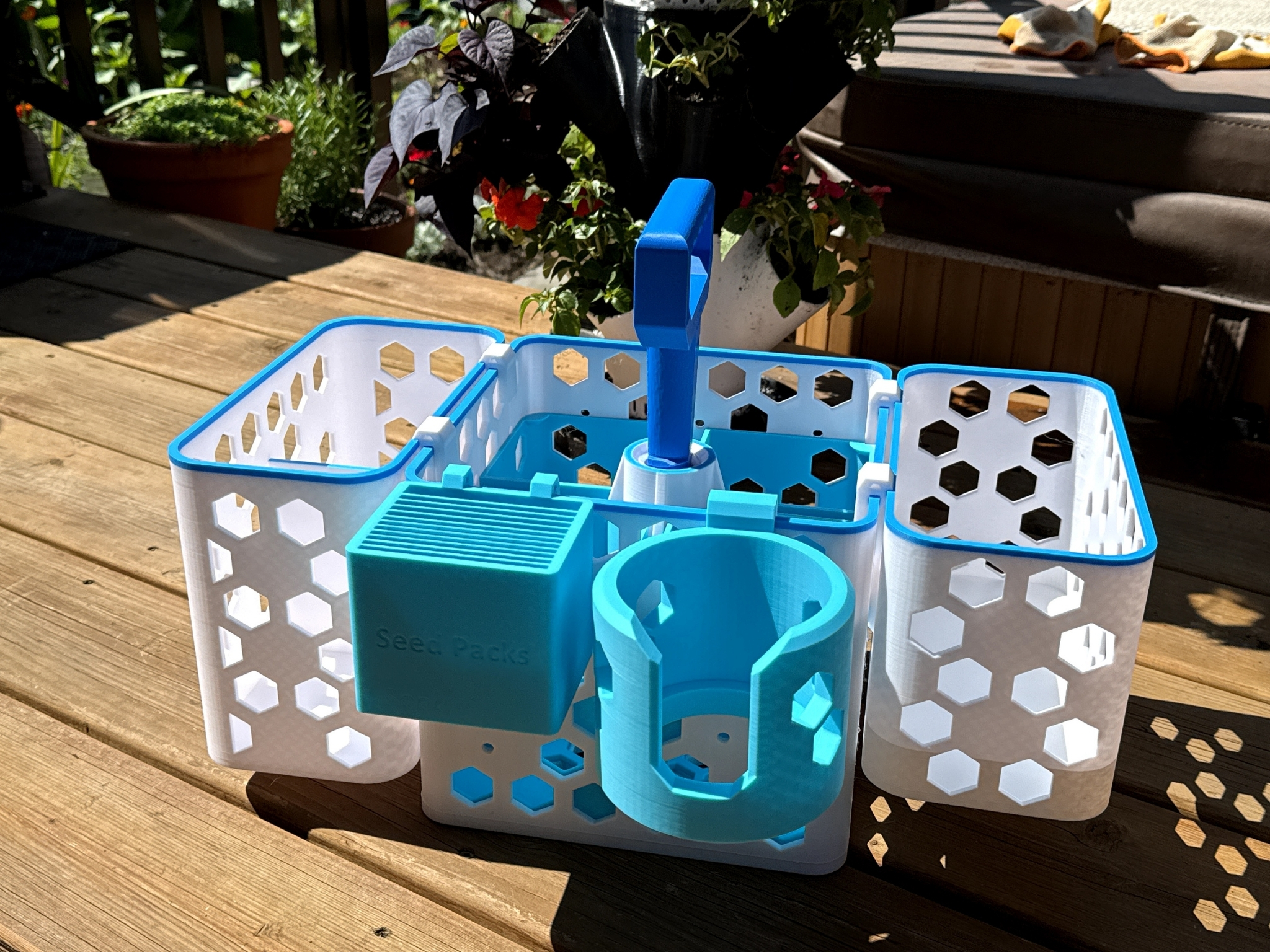 2 Multi-compartment seed storage organizer in vibrant blue and white, designed for easy access to seed packs, featuring hexagonal cutouts and a convenient handle, placed on a wooden deck surrounded by plants.
