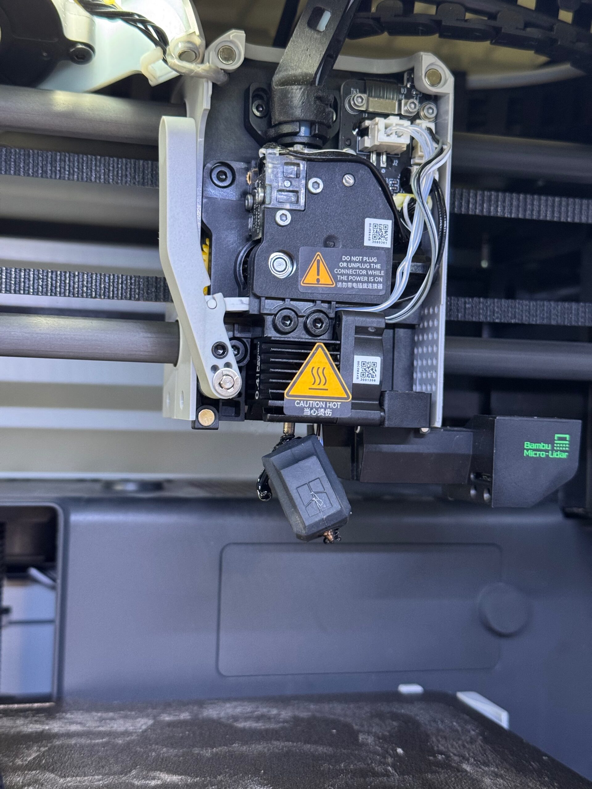 E9CB72D4-7FA0-4CBA-8582-00A5FA370434_L0_001-2025-05-31, 8_34_43 PM Close-up of a 3D printer's hotend assembly showcasing components such as the nozzle, wiring, and safety warning labels. The image highlights the intricate design and features of the printer's extruder mechanism, including a Micro-Lidar sensor for enhanced precision.